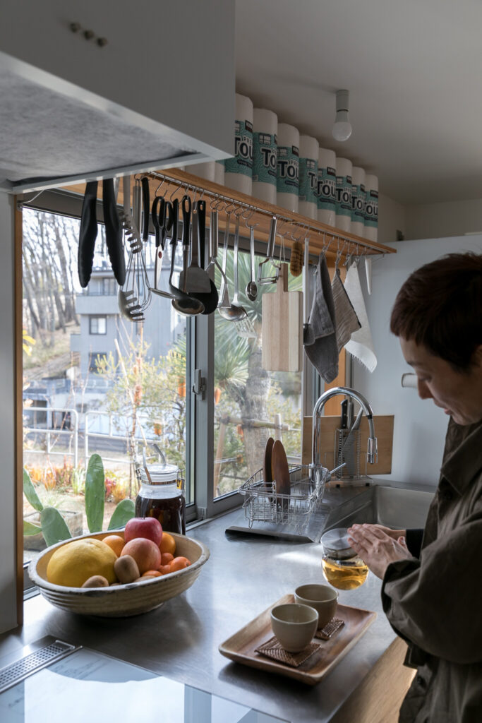 窓越しにリビングが見える明るいキッチン。料理道具をかけている棚板はDIYで。